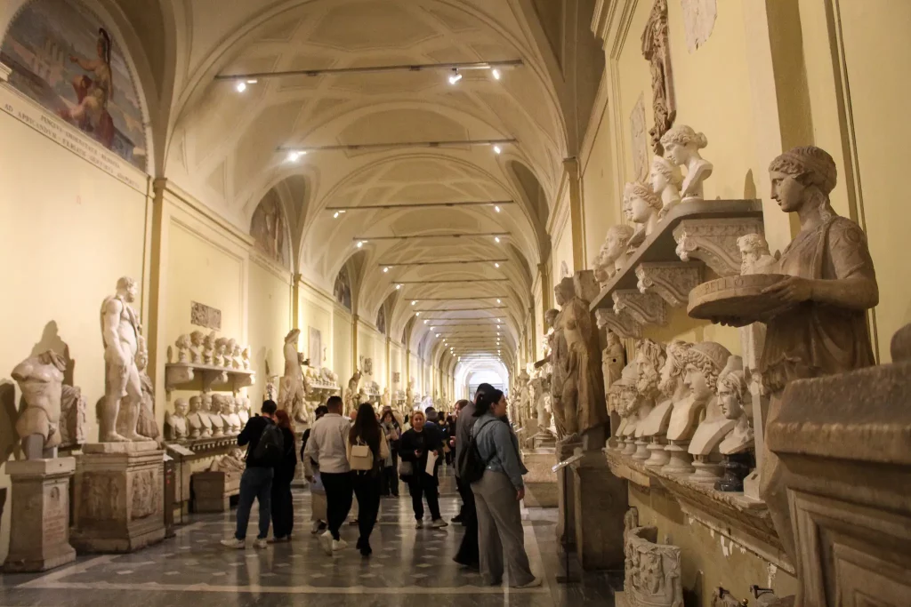 Visitors walking through the Hall of the Muses in the Vatican Museums, surrounded by classical Roman sculptures and marble busts